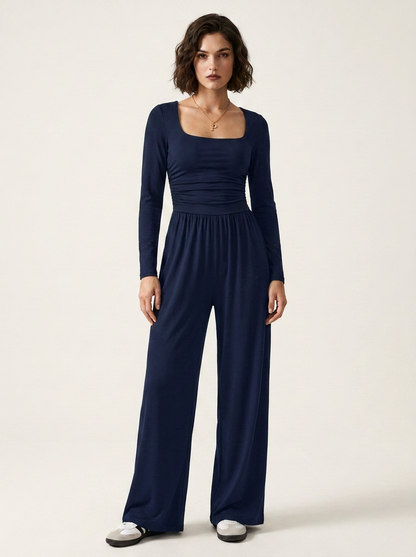 A woman modeling a navy blue long-sleeve jumpsuit with a square neckline and wide legs, standing against a plain off-white studio background.