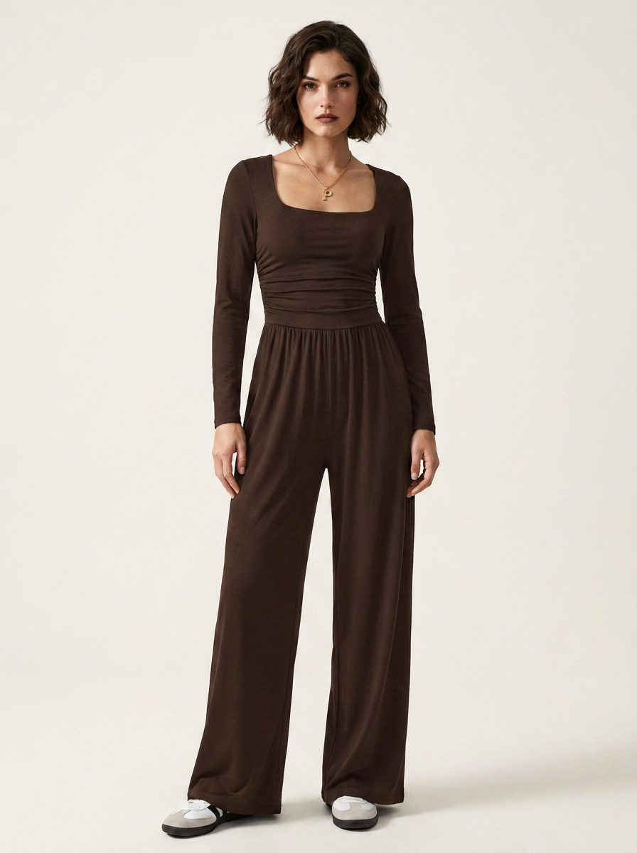 A woman modeling a dark brown long-sleeve jumpsuit with a square neckline and wide-leg pants, standing against a plain light beige studio background.