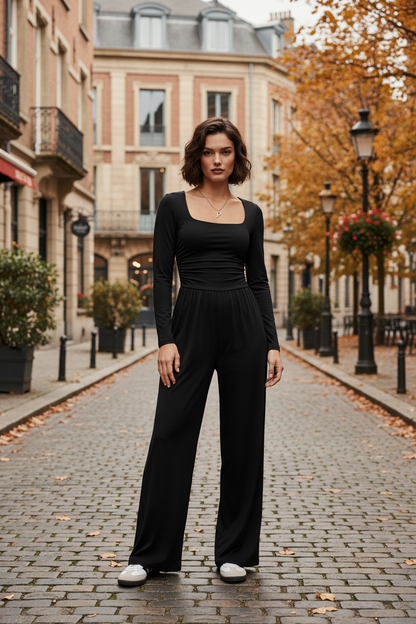 A woman stands on a cobblestone street wearing a black long-sleeve jumpsuit with a square neckline and wide legs, paired with white sneakers. The background shows a softly blurred urban street scene with buildings, trees, and lampposts.