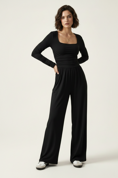 A woman modeling a black long-sleeve jumpsuit with a square neckline, gathered waist, and wide legs, photographed against a plain light beige studio background.