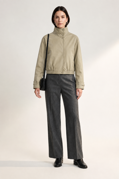 A model stands against a light studio background wearing a cream high-collar jacket, dark gray wide-leg trousers, black shoes, and carrying a small black shoulder bag.