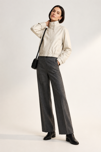 A model stands against a light studio background wearing a cream high-collar jacket, dark gray wide-leg trousers, black shoes, and carrying a small black shoulder bag.