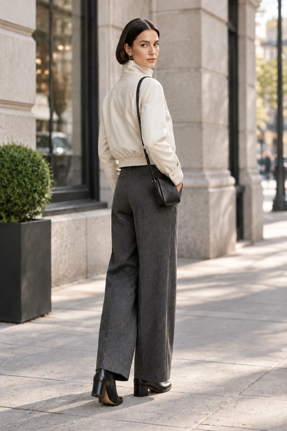 A woman walking on a city sidewalk wearing a cream zip-front jacket, dark gray wide-leg trousers, black ankle boots, and carrying a black shoulder bag.