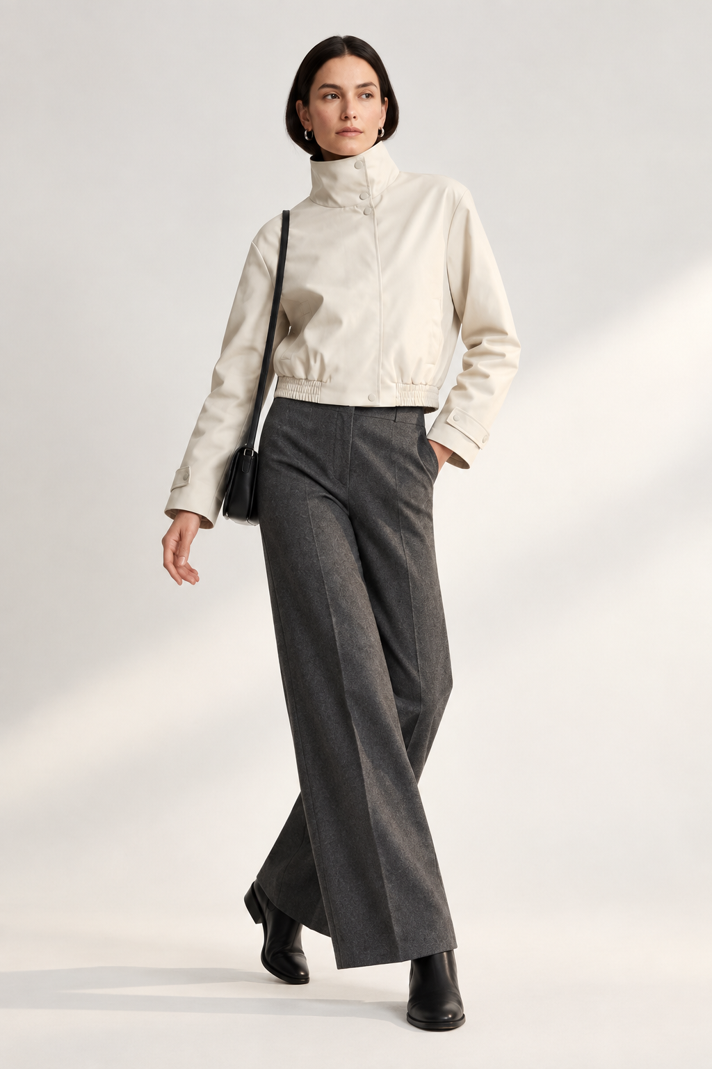 A model stands against a light studio background wearing a cream high-collar jacket, dark gray wide-leg trousers, black shoes, and carrying a small black shoulder bag.