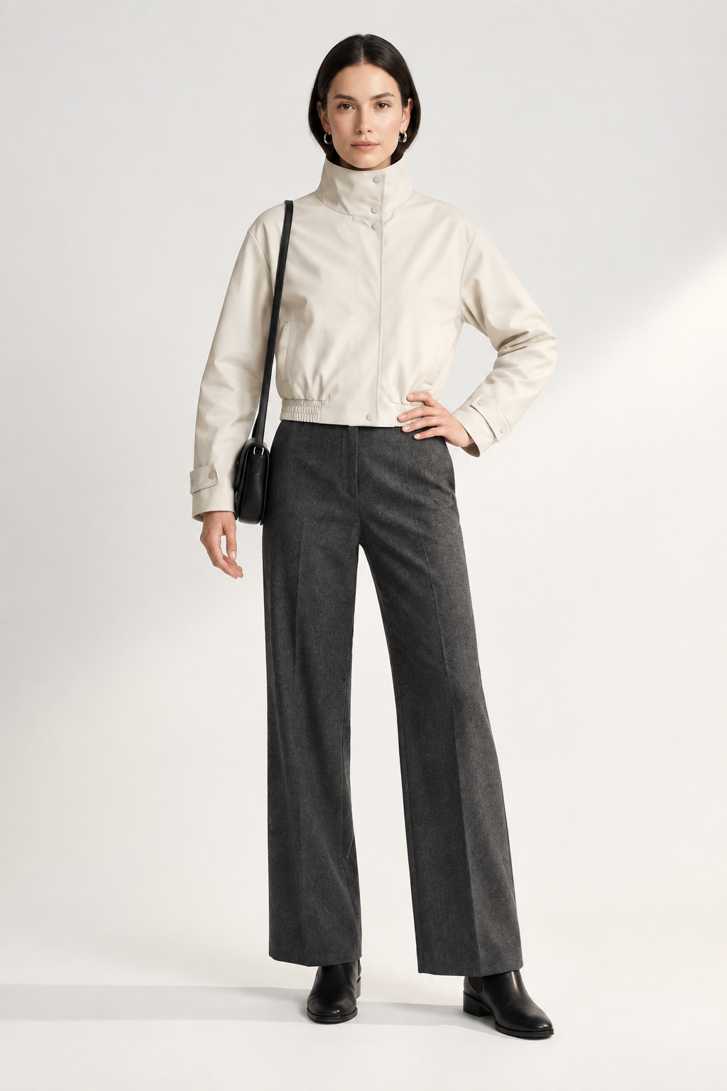 A model stands against a light studio background wearing a cream high-collar jacket, dark gray wide-leg trousers, black shoes, and carrying a small black shoulder bag.