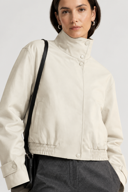 A model stands against a light studio background wearing a cream high-collar jacket, dark gray wide-leg trousers, black shoes, and carrying a small black shoulder bag.