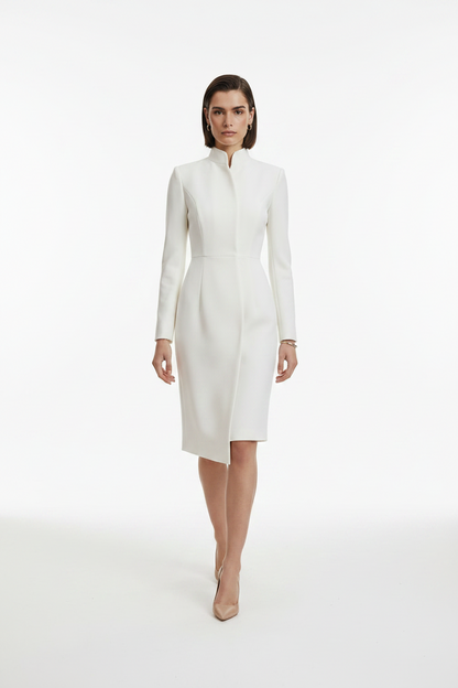 A model wearing a structured white long-sleeve midi dress with a high mock neck and asymmetrical hem, paired with beige pointed-toe heels, against a seamless white studio background.