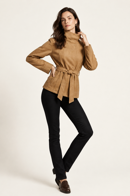 A woman modeling a tan suede belted jacket with a high mock neck, paired with black pants, against a plain off-white studio background.