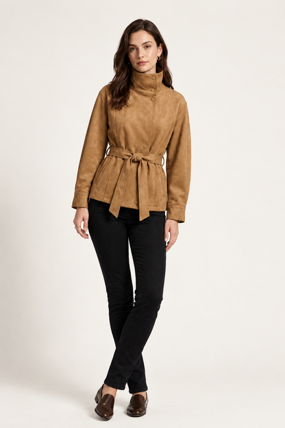 A woman modeling a tan suede belted jacket with a high mock neck, paired with black pants, against a plain off-white studio background.