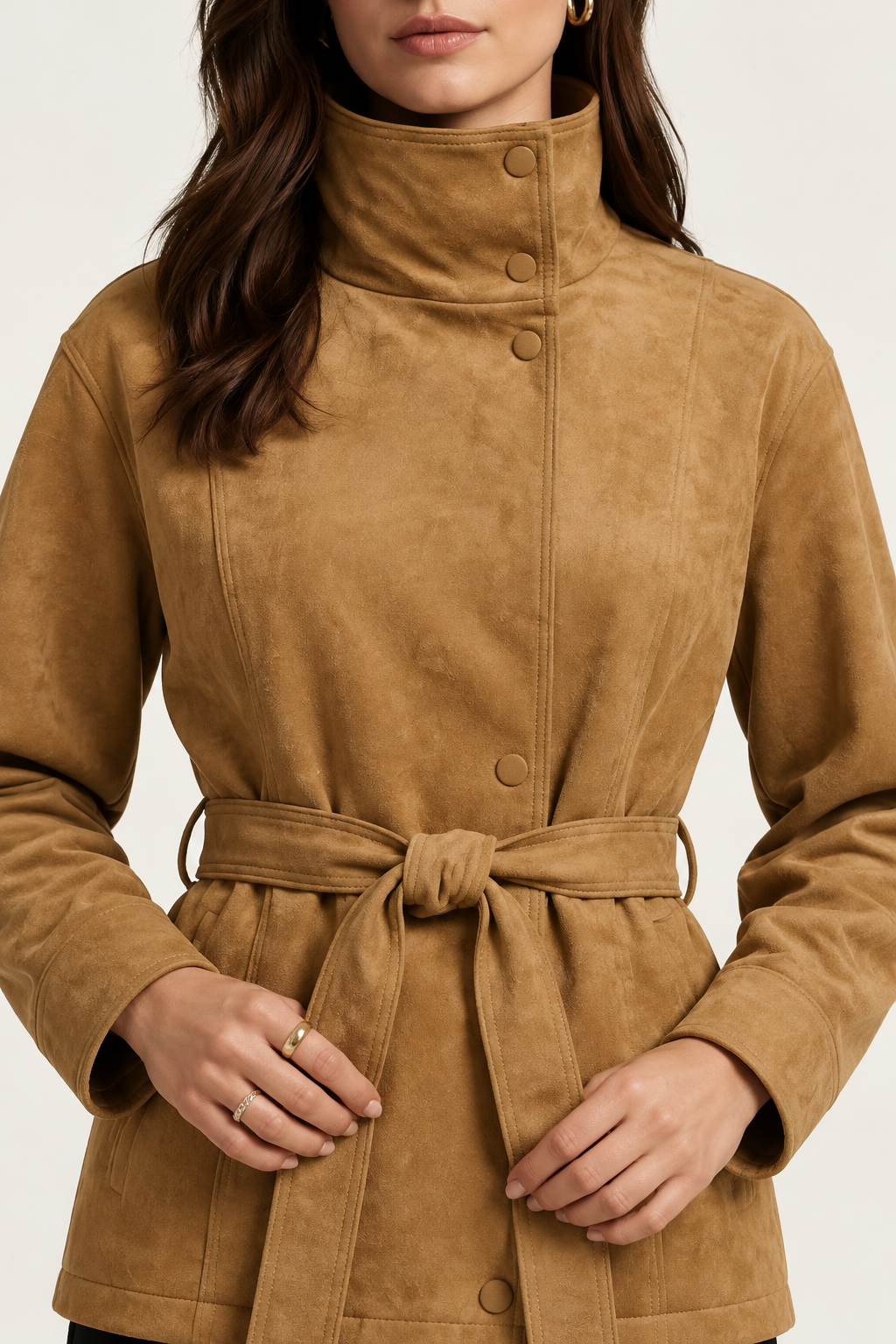 A woman modeling a tan suede belted jacket with a high mock neck, paired with black pants, against a plain off-white studio background.