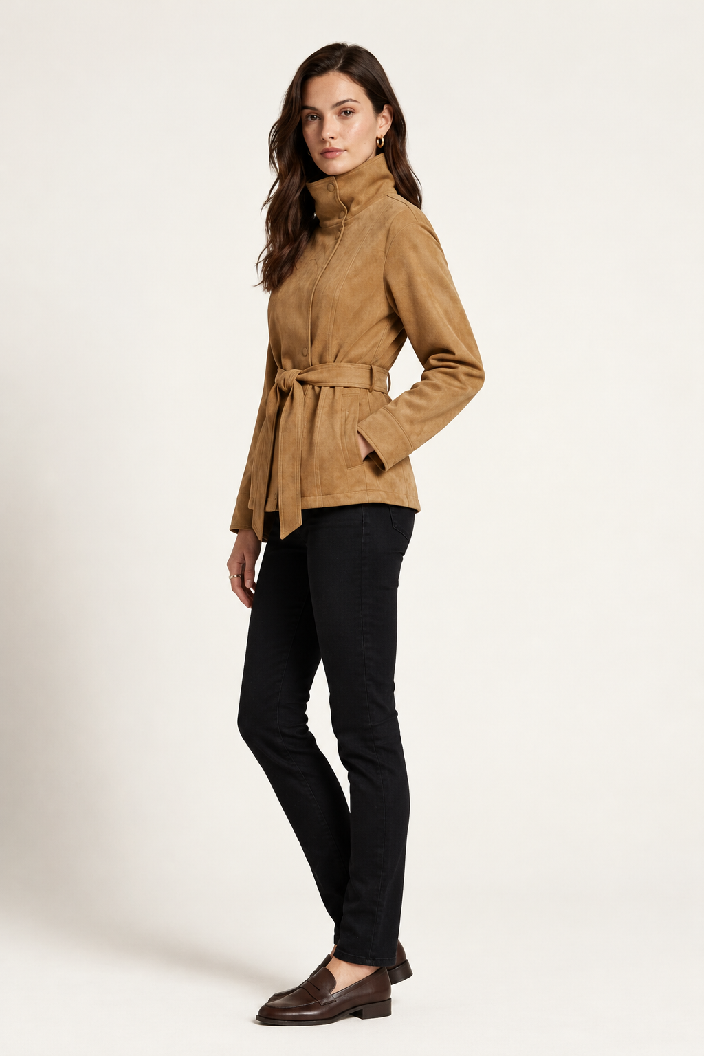 A woman modeling a tan suede belted jacket with a high mock neck, paired with black pants, against a plain off-white studio background.
