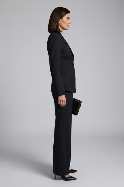 A woman modeling a tailored black two-piece suit with a structured blazer and wide-leg trousers, holding a black clutch against a light gray studio background.
