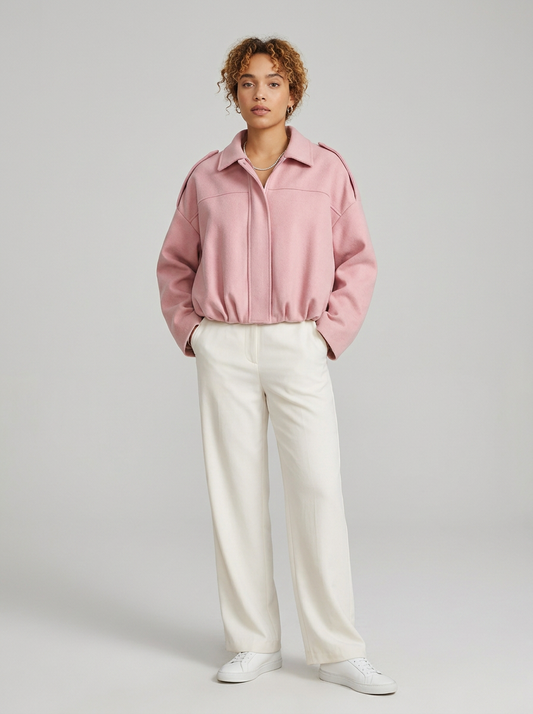 A model stands against a light gray studio background wearing a dusty pink cropped jacket with shoulder epaulets, off-white wide-leg trousers, and white sneakers.