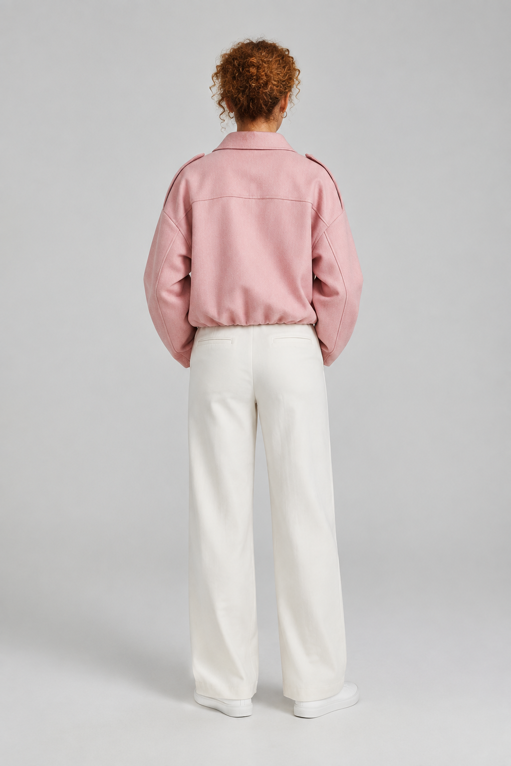 A model viewed from the back wearing a light pink cropped jacket, white wide-leg trousers, and white sneakers, set against a neutral light gray studio background.