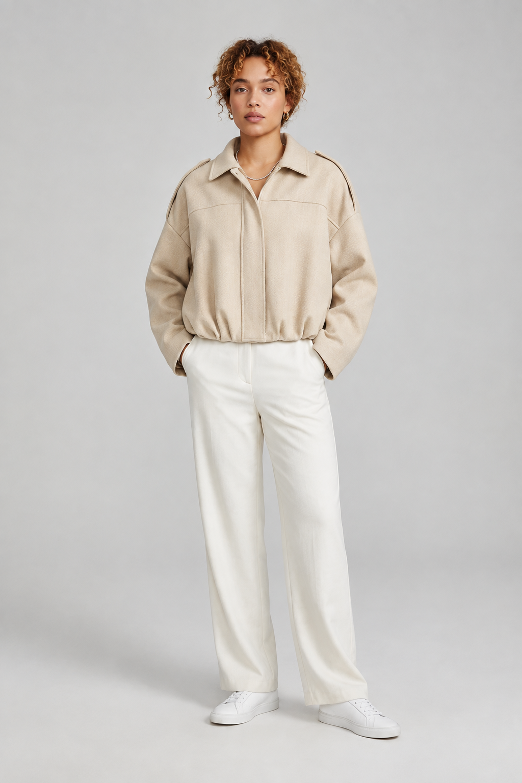 A model stands against a light gray studio background wearing a dusty pink cropped jacket with shoulder epaulets, off-white wide-leg trousers, and white sneakers.