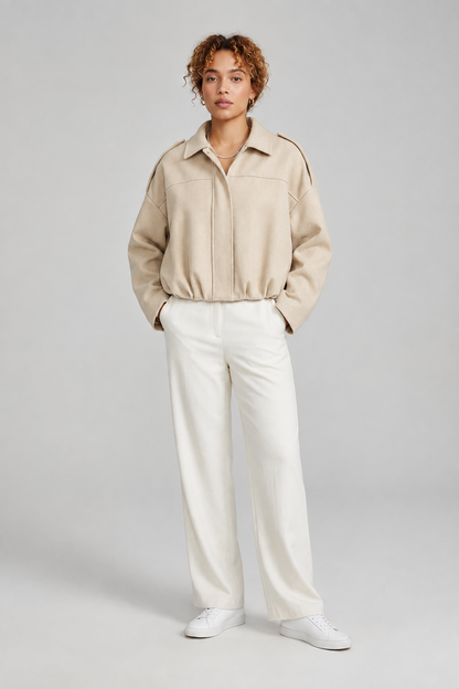 A model stands against a light gray studio background wearing a dusty pink cropped jacket with shoulder epaulets, off-white wide-leg trousers, and white sneakers.