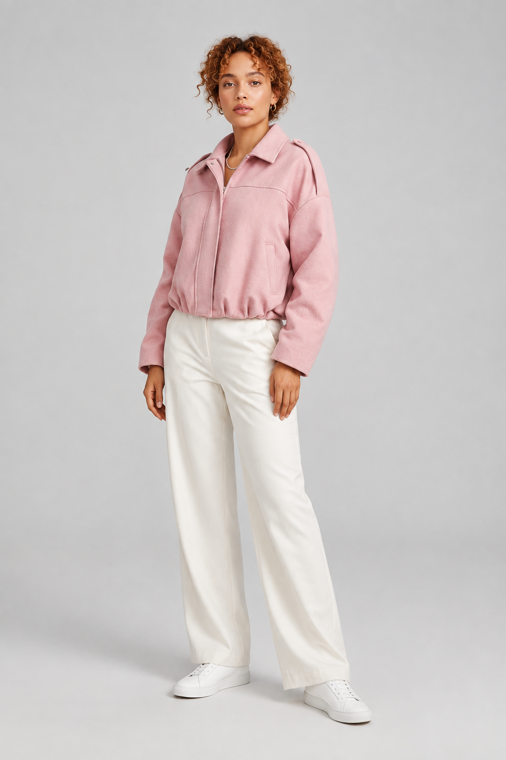 A model stands against a light gray studio background wearing a light pink zip-front jacket, white wide-leg trousers, and white sneakers.