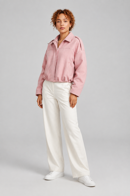 A model stands against a light gray studio background wearing a light pink zip-front jacket, white wide-leg trousers, and white sneakers.