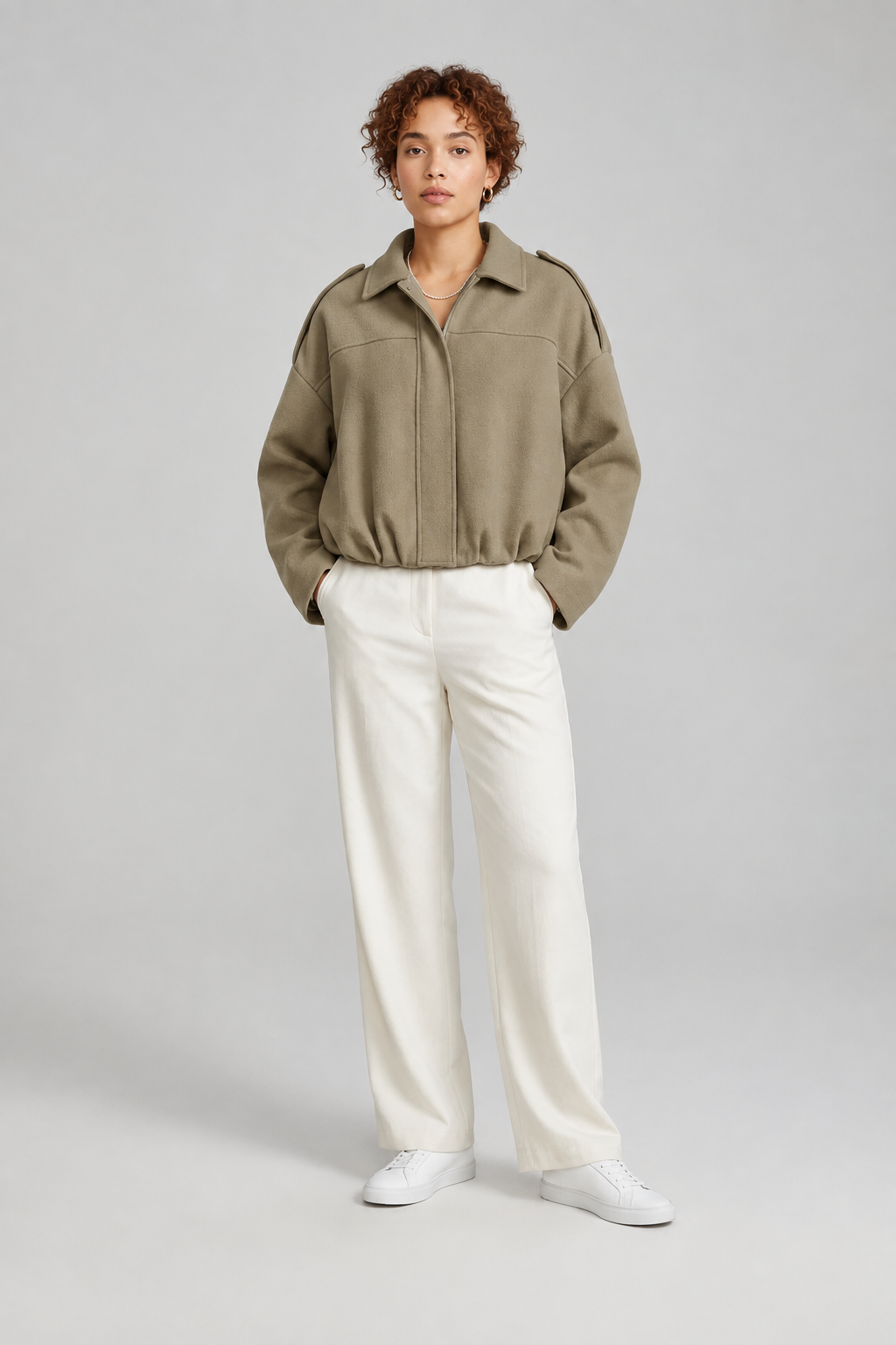 A model stands against a light gray studio background wearing a dusty pink cropped jacket with shoulder epaulets, off-white wide-leg trousers, and white sneakers.