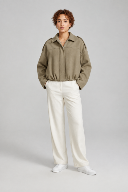 A model stands against a light gray studio background wearing a dusty pink cropped jacket with shoulder epaulets, off-white wide-leg trousers, and white sneakers.