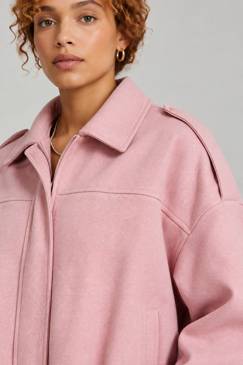 A close-up of a model wearing a dusty pink textured jacket with a wide collar and shoulder epaulets, set against a solid light gray background.