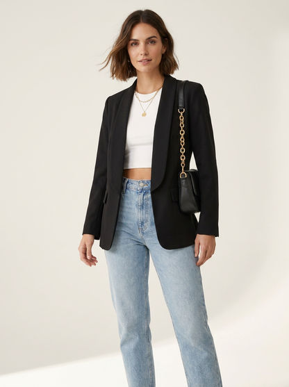 A woman modeling a black blazer over a white crop top and light blue jeans, carrying a black shoulder bag with a gold chain strap against a plain light beige studio background.