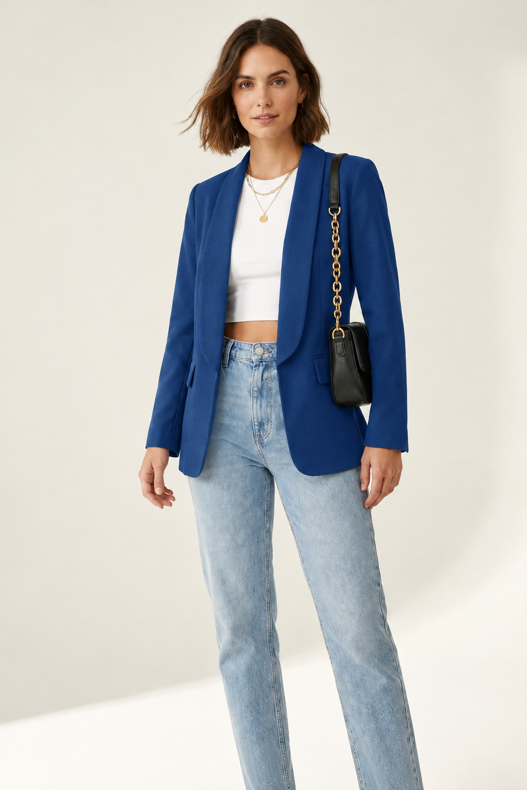 A woman modeling a black blazer over a white crop top and light blue jeans, carrying a black shoulder bag with a gold chain strap against a plain light beige studio background.