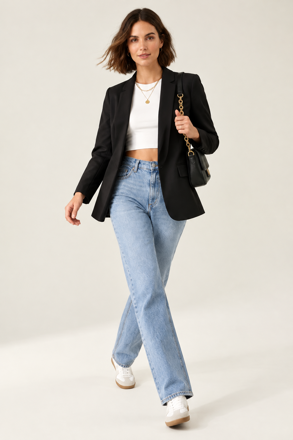 A woman modeling a black blazer over a white crop top and light blue jeans, carrying a black shoulder bag with a gold chain strap against a plain light beige studio background.
