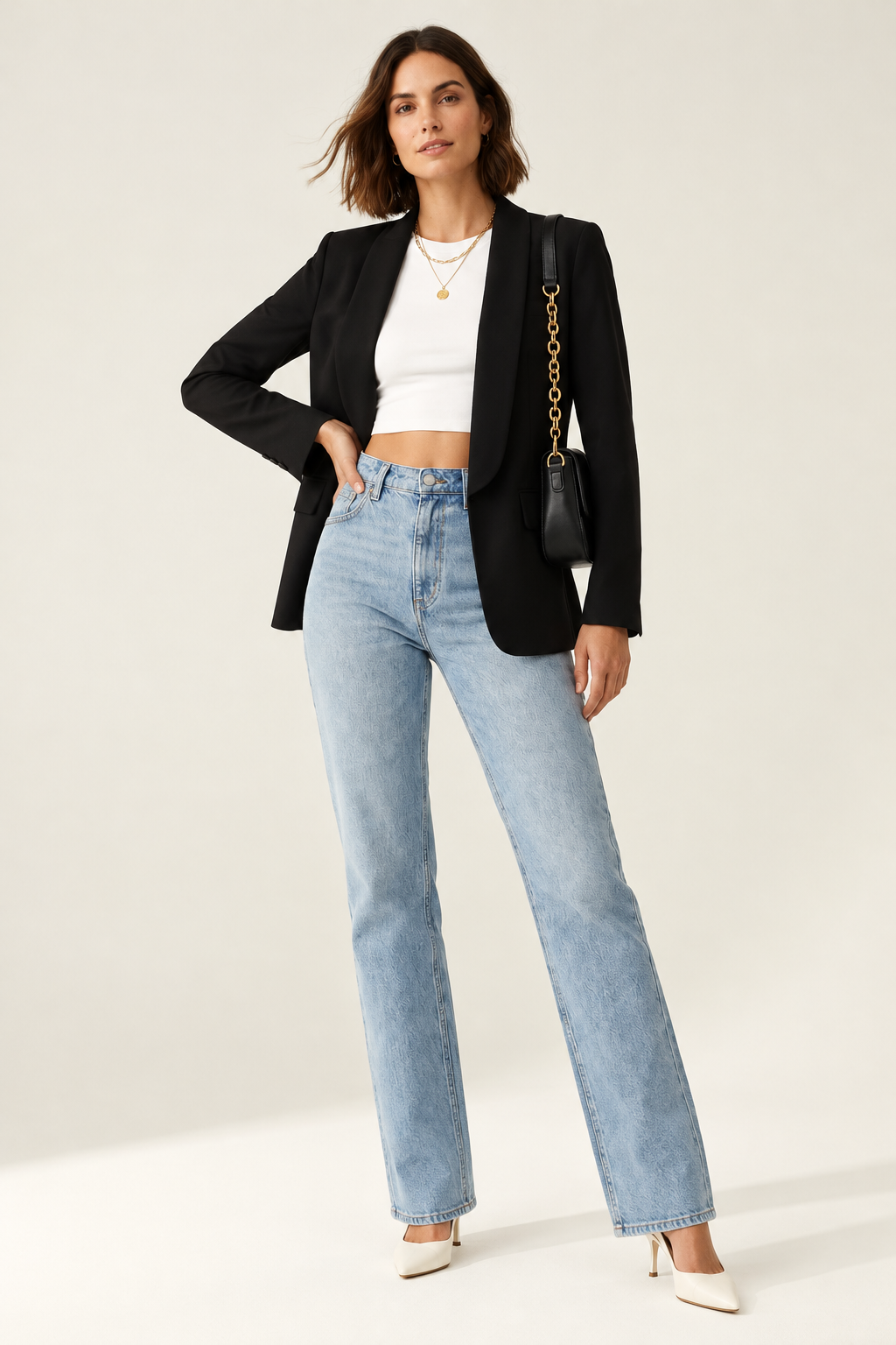 A woman modeling a black blazer over a white crop top and light blue jeans, carrying a black shoulder bag with a gold chain strap against a plain light beige studio background.