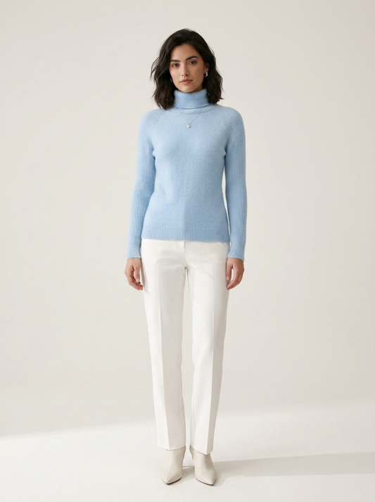 A woman stands against a plain light beige studio background wearing a light blue ribbed turtleneck sweater, white trousers, and white pointed-toe heels.