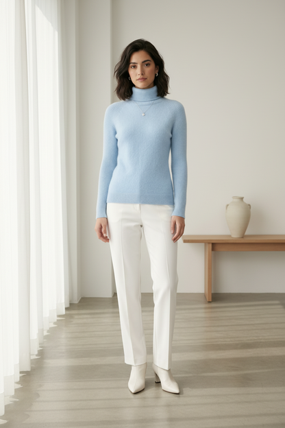 A woman stands against a plain light beige studio background wearing a light blue ribbed turtleneck sweater, white trousers, and white pointed-toe heels.