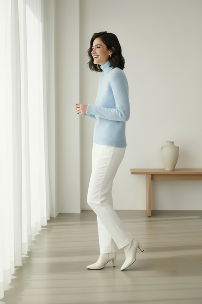 A woman stands in profile in a bright, minimalist room wearing a light blue turtleneck sweater, white pants, and white ankle boots. A small wooden table with a vase is visible in the background near a window with sheer curtains.