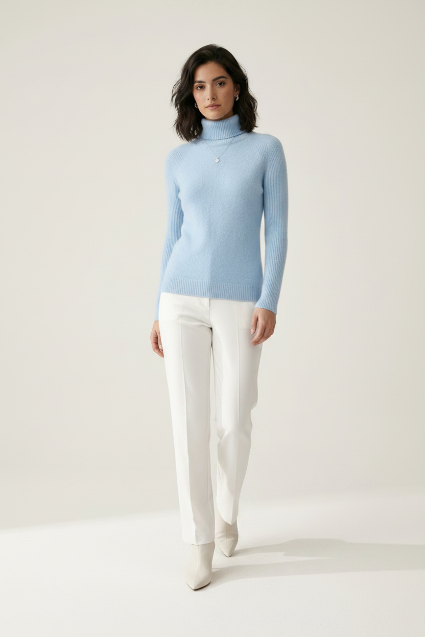 A woman stands against a plain light beige studio background wearing a light blue ribbed turtleneck sweater, white trousers, and white pointed-toe heels.