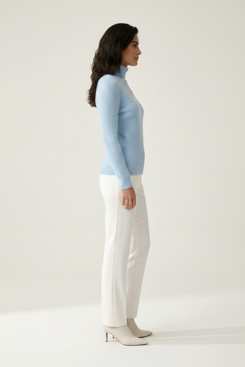 A woman stands against a plain light beige studio background wearing a light blue ribbed turtleneck sweater, white trousers, and white pointed-toe heels.