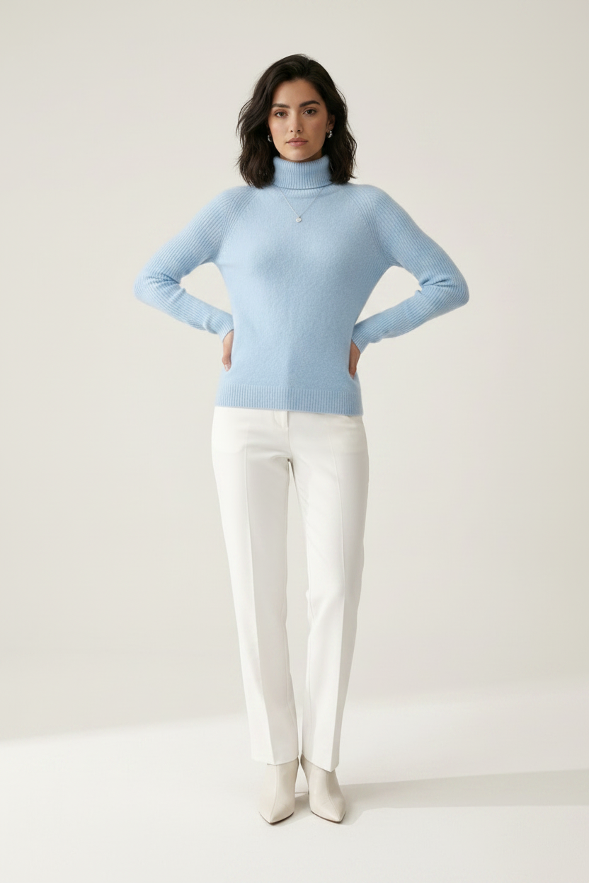 A woman stands against a plain light beige studio background wearing a light blue ribbed turtleneck sweater, white trousers, and white pointed-toe heels.