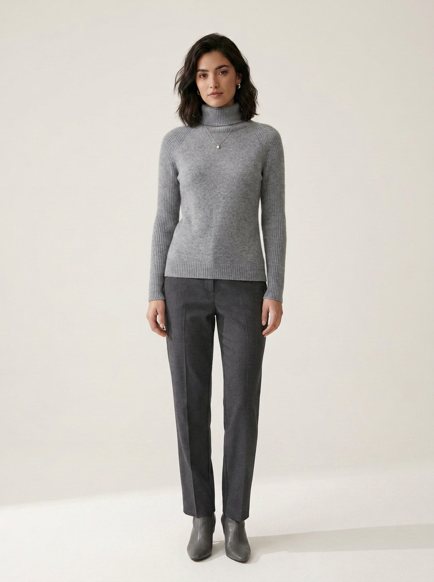 A woman stands against a plain light beige studio background wearing a heather gray ribbed turtleneck sweater, dark charcoal gray trousers, and gray ankle boots.