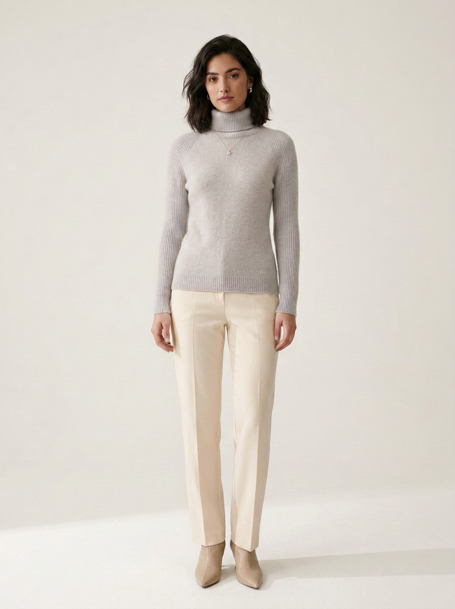 A woman stands against a plain light beige studio background wearing a light gray ribbed turtleneck sweater, cream-colored straight-leg trousers, and beige pointed-toe heels.