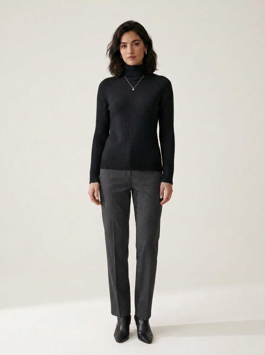 A woman stands against a plain light beige studio background wearing a black long-sleeve mock-neck sweater, dark gray straight-leg trousers, and black ankle boots.