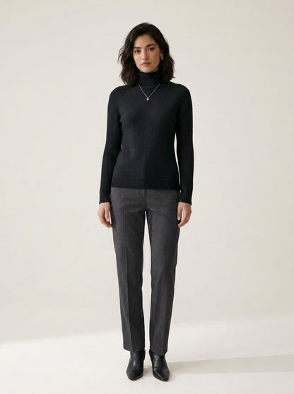 A woman stands against a plain light beige studio background wearing a black long-sleeve mock-neck sweater, dark gray straight-leg trousers, and black ankle boots.