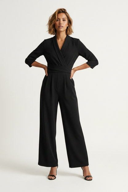 A woman modeling a black wrap-style jumpsuit with three-quarter sleeves, a self-tie waist belt, and wide legs, paired with black heeled sandals against a plain light background.