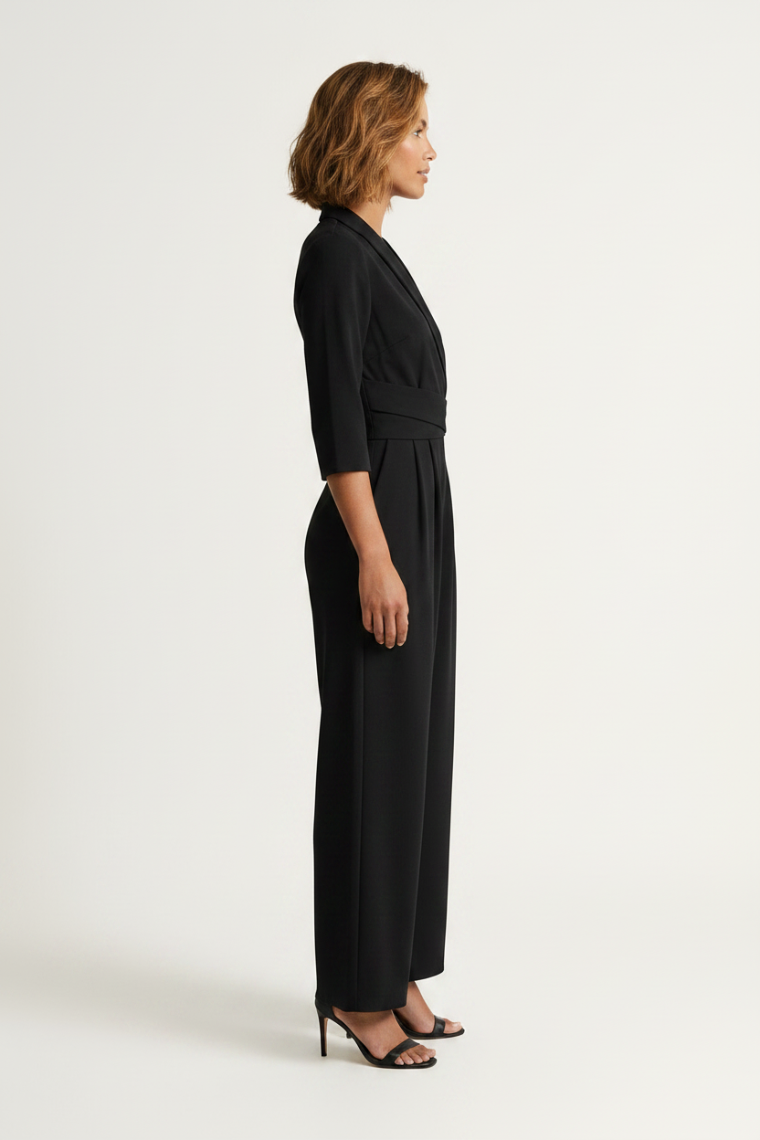 A woman modeling a black wrap-style jumpsuit with three-quarter sleeves, a self-tie waist belt, and wide legs, paired with black heeled sandals against a plain light background.