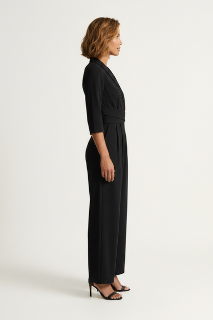 A woman modeling a black wrap-style jumpsuit with three-quarter sleeves, a self-tie waist belt, and wide legs, paired with black heeled sandals against a plain light background.