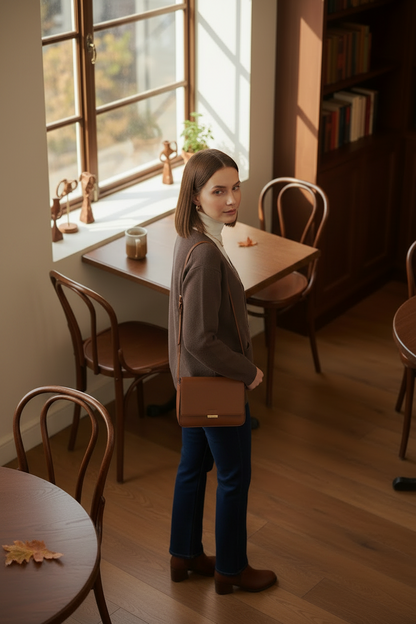 A woman stands in a warm, sunlit room with wooden floors and bookshelves, wearing a brown cardigan over a white turtleneck, dark blue jeans, and brown ankle boots.
