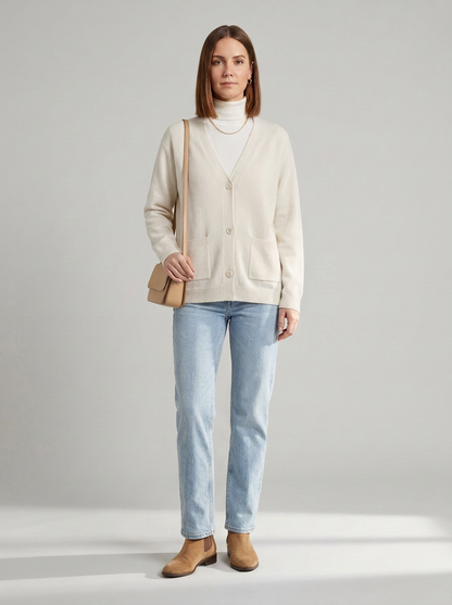 A woman stands against a light gray studio background wearing a cream cardigan over a white turtleneck, light blue jeans, and tan ankle boots, while carrying a small tan shoulder bag.