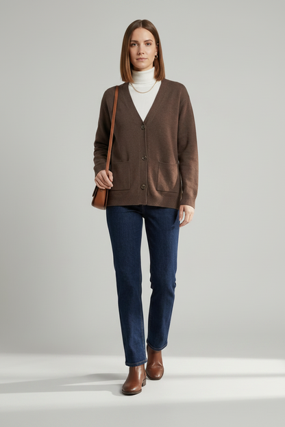 A woman stands against a light gray studio background wearing a brown button-front cardigan over a white turtleneck, dark blue jeans, and brown leather ankle boots, with a small brown shoulder bag.