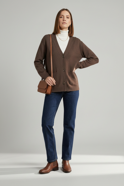 A woman stands against a light gray studio background wearing a brown button-front cardigan over a white turtleneck, dark blue jeans, and brown leather ankle boots, with a small brown shoulder bag.