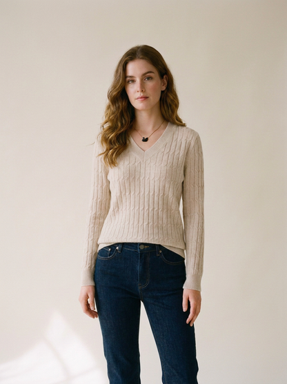 A woman modeling a beige cable-knit V-neck sweater with dark blue jeans, standing against a plain light beige studio background.