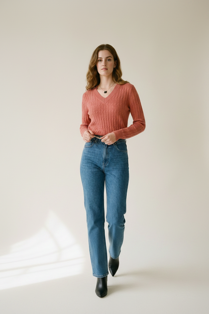 A woman modeling a ribbed long-sleeve V-neck sweater in a dusty rose pink color, paired with high-waisted blue jeans, against a plain beige studio background.