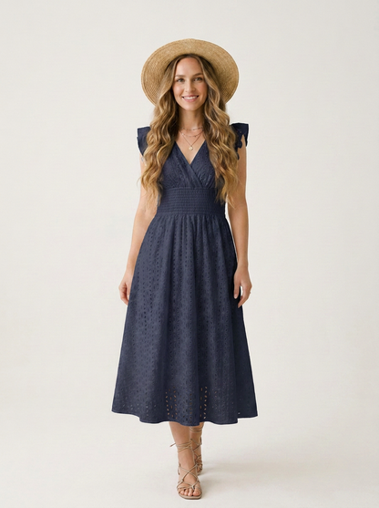 A woman modeling a sleeveless navy blue eyelet midi dress with a V-neckline and cap sleeves, styled with a straw hat and beige strappy sandals against a plain light background.