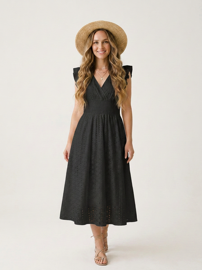 A woman modeling a sleeveless black midi dress with a V-neckline and flared skirt, styled with a wide-brimmed straw hat and strappy high-heeled sandals against a plain light background.